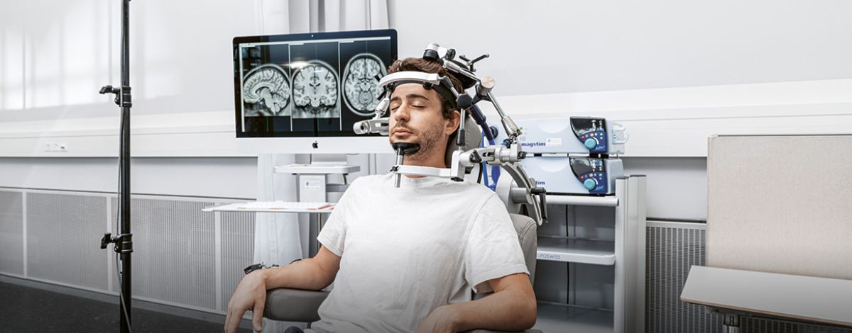 Man seated with eyes closed as a TMS coil is held against his head, with brain scans on a monitor behind him