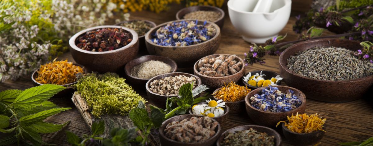 Wooden bowls of dried herbs, flowers and spices on a wooden table beside a white mortar and pestle