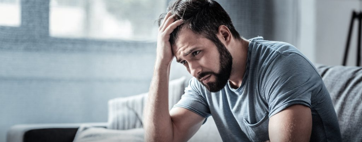 Bearded man in a striped t-shirt sitting on a sofa with one hand on his head and a worried look