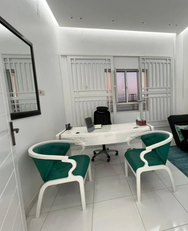 Consulting room with a white desk, a black office chair and two green upholstered chairs facing it, window behind