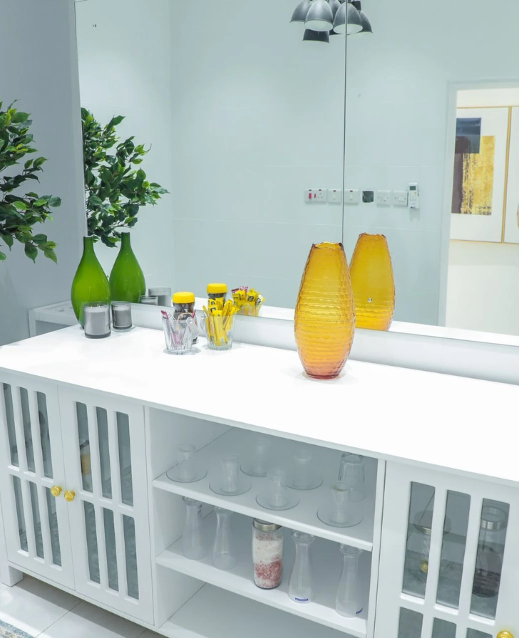 White cabinet with glass-fronted doors, amber and green vases, drinking glasses and sachets, in front of a wall mirror