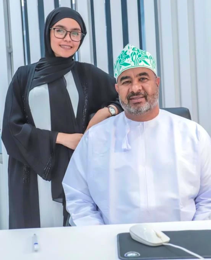 Man in a white robe and green patterned cap seated at a desk, smiling, with a woman in a black abaya and glasses beside him