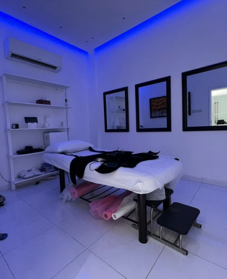 Treatment room lit by blue ceiling lights, with a padded couch, a black device with cables on it, wall mirrors and shelves