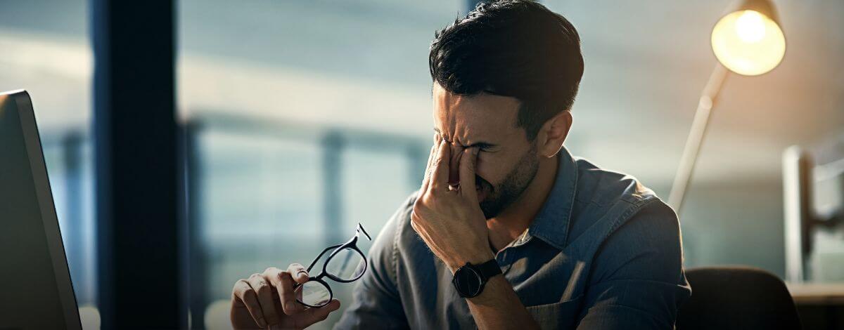 Man at a desk pressing his fingers to his eyes with one hand and holding glasses in the other, lamp glowing behind