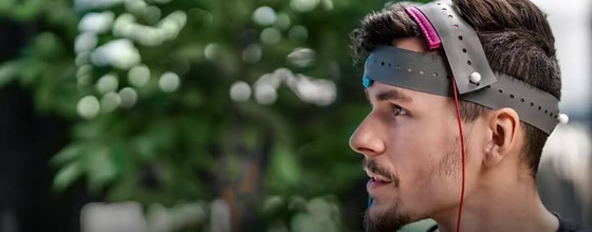 Man in profile wearing a grey perforated headband with electrodes and a red wire, with blurred greenery behind