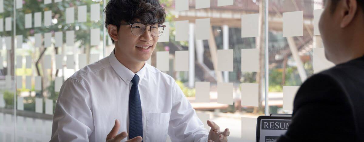 Hussain K., University Student, in glasses, white shirt and dark tie, speaking with someone across a table