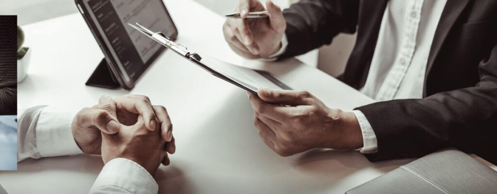Two people in suits at a table reviewing a form on a clipboard
