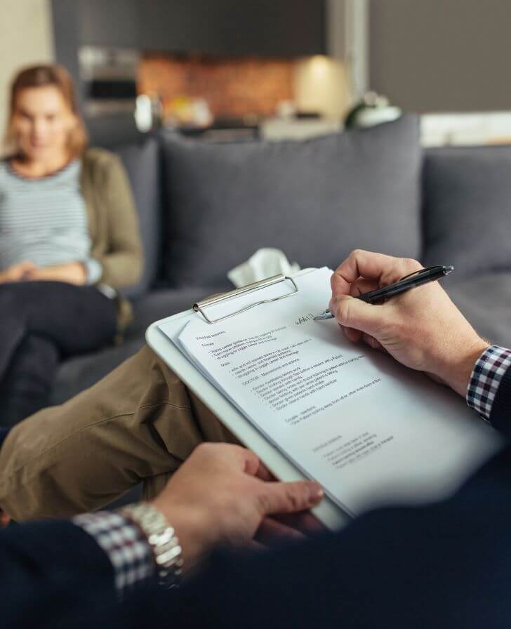 Person writing notes on a clipboard in the foreground, with a woman sitting on a grey sofa out of focus behind