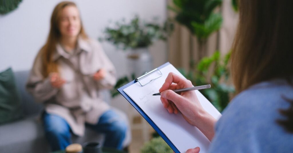 Clinician taking notes on a clipboard while a woman sits on a sofa