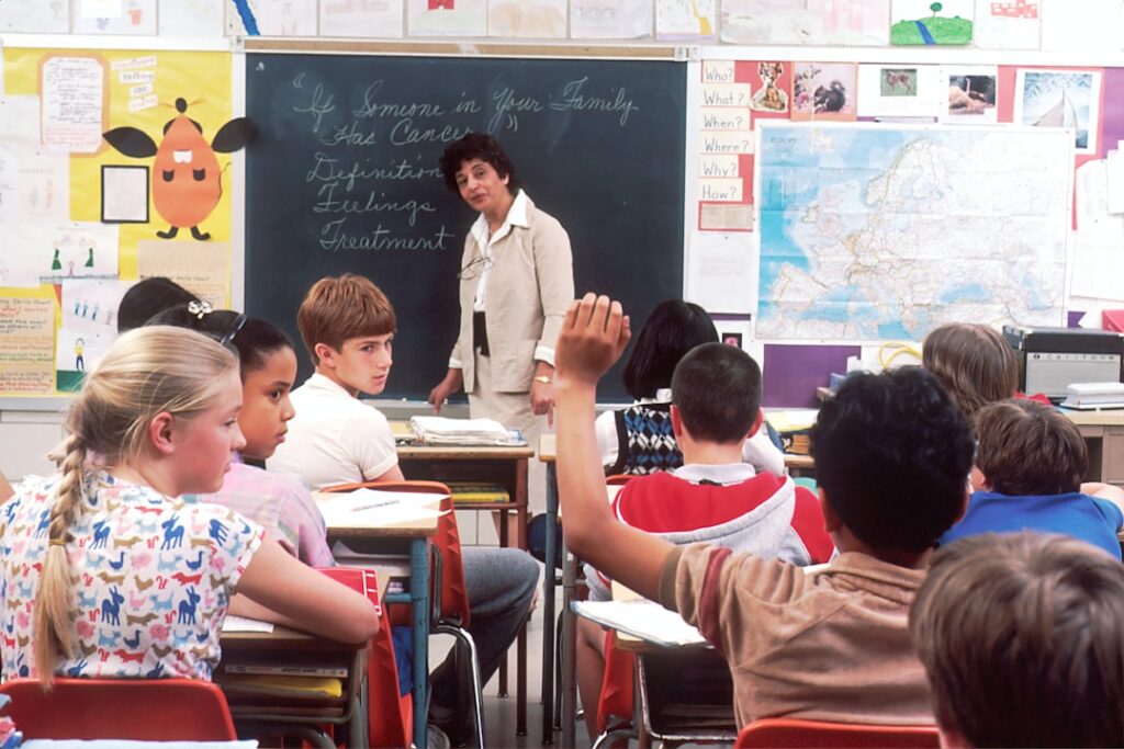 Teacher at a blackboard with a classroom of children, one with a raised hand