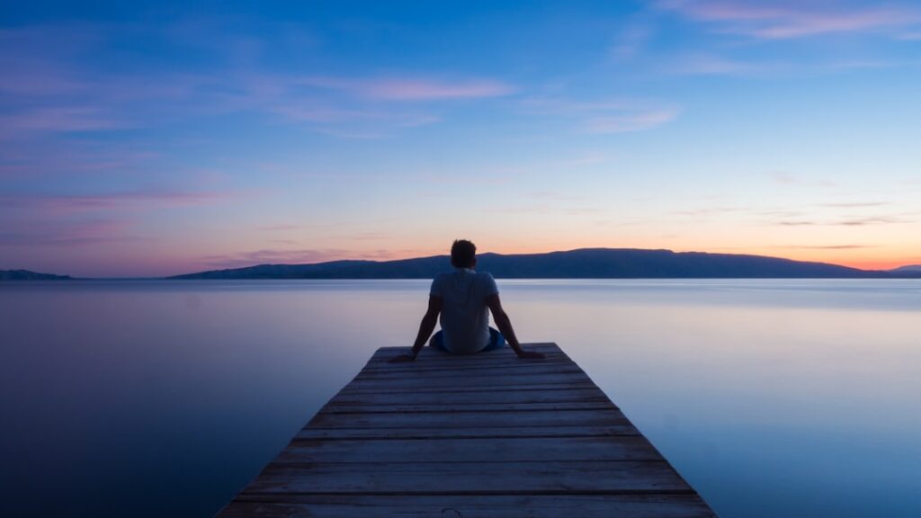 Person sitting at the end of a wooden jetty looking over still water at sunset