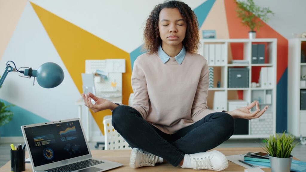 Woman meditating cross-legged on a desk beside a laptop
